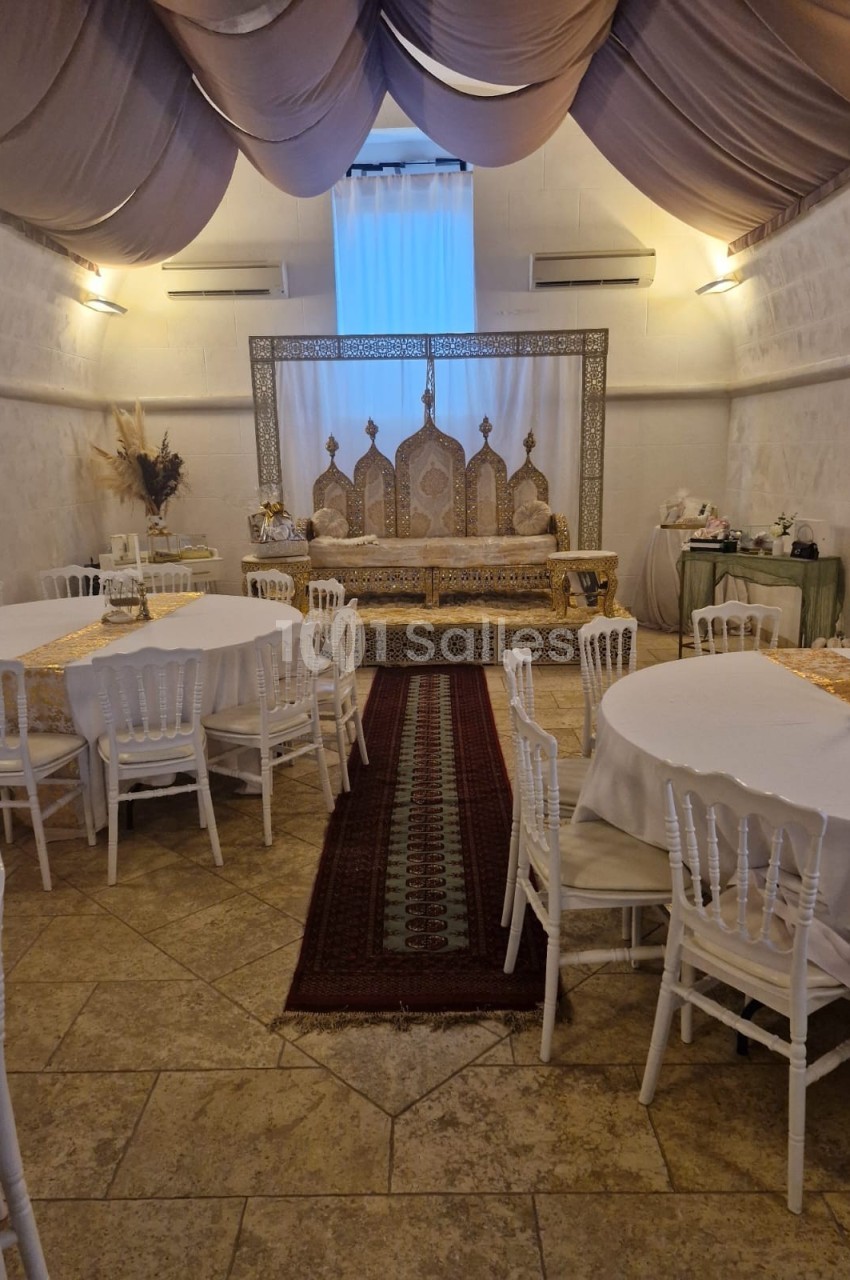 Salle décorée pour un événement avec tables rondes, chaises blanches et une estrade ornée de motifs orientaux.