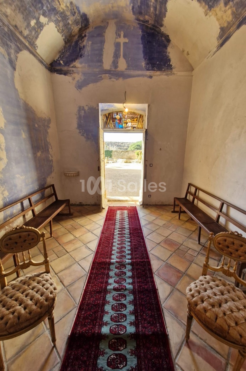 Intérieur d'une petite chapelle avec un tapis rouge central, des bancs en bois et une porte ouverte sur un paysage lumineux.