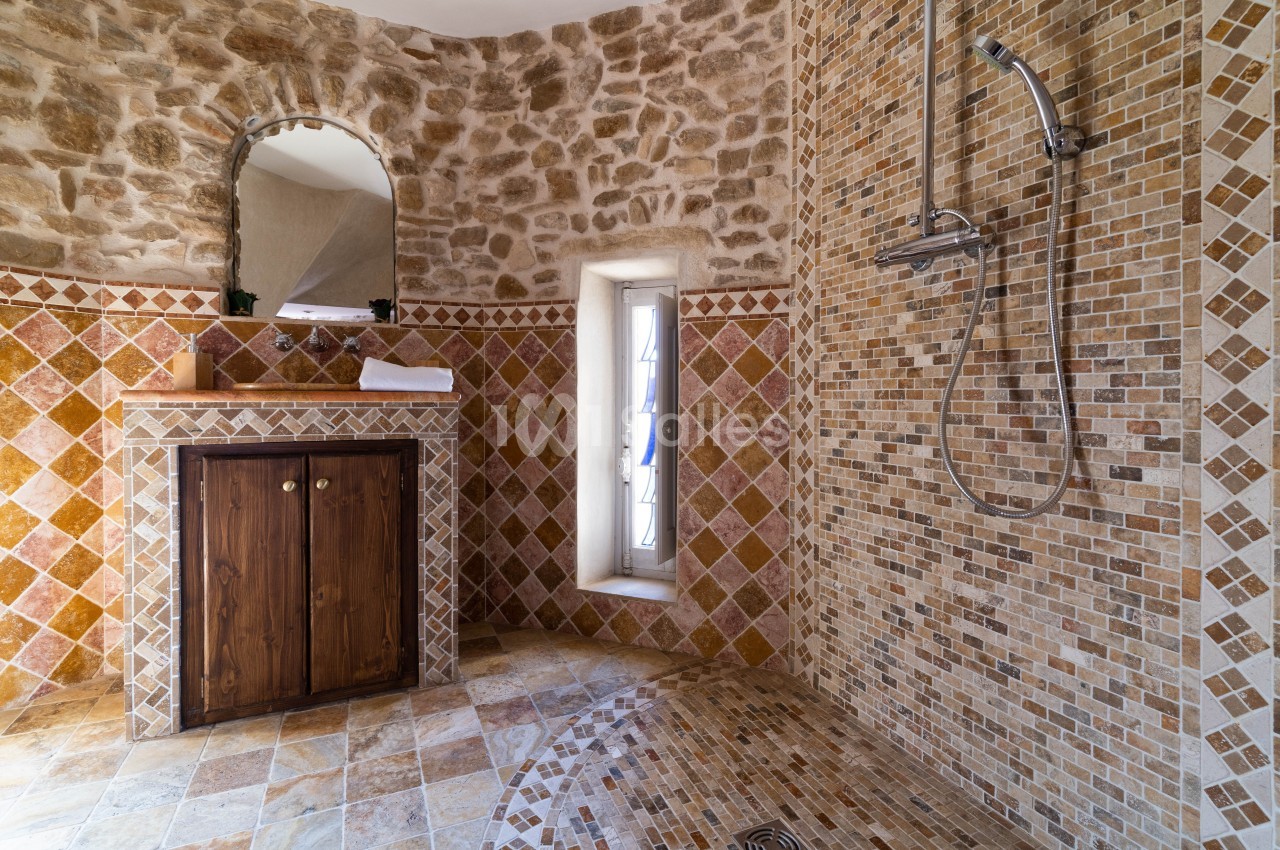 Salle de bain avec murs en pierre et mosaïque, douche à l'italienne et meuble en bois sous un miroir.