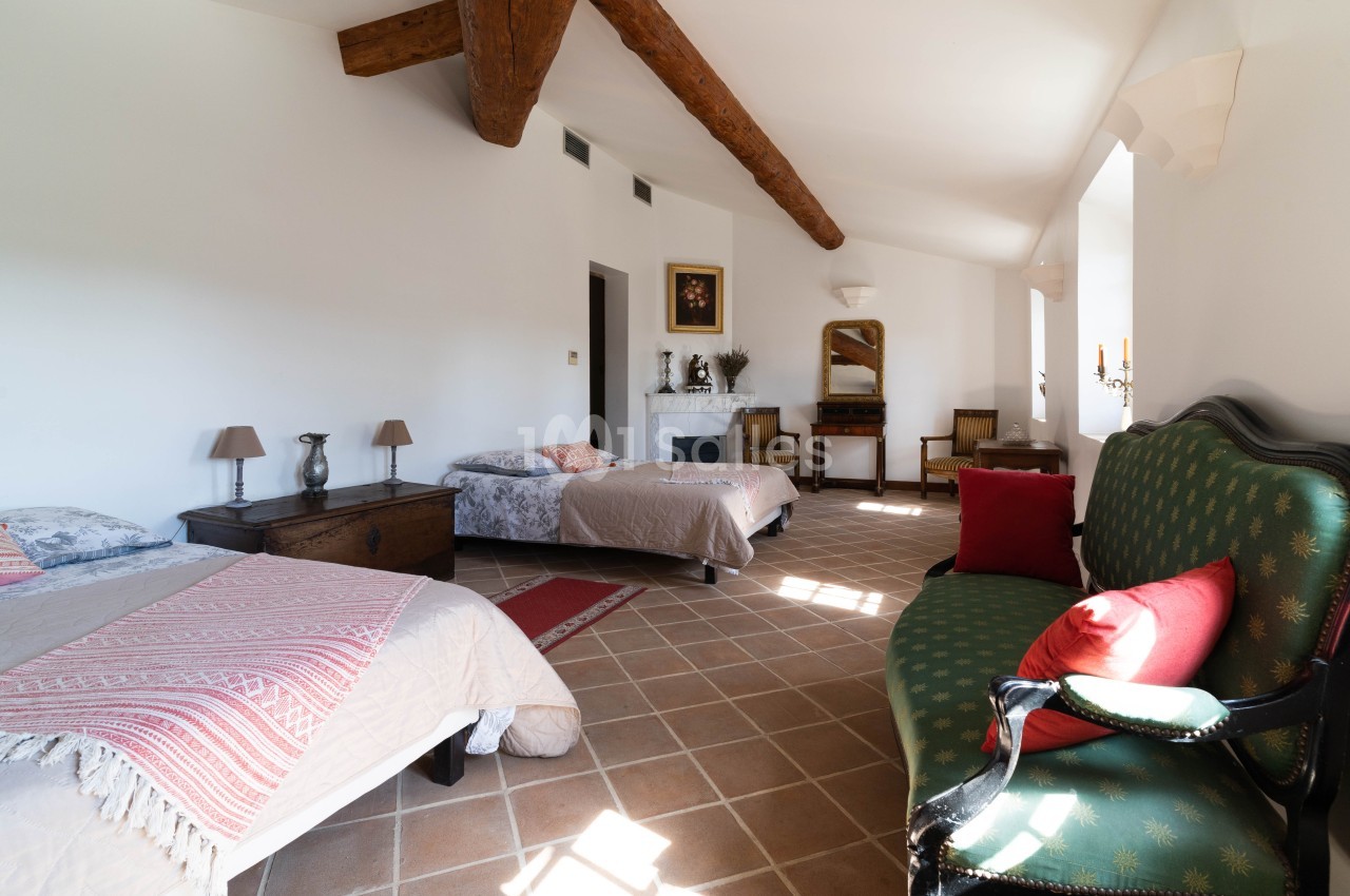 Chambre lumineuse avec deux lits simples, canapé, poutres apparentes, sol en carreaux et décoration classique.