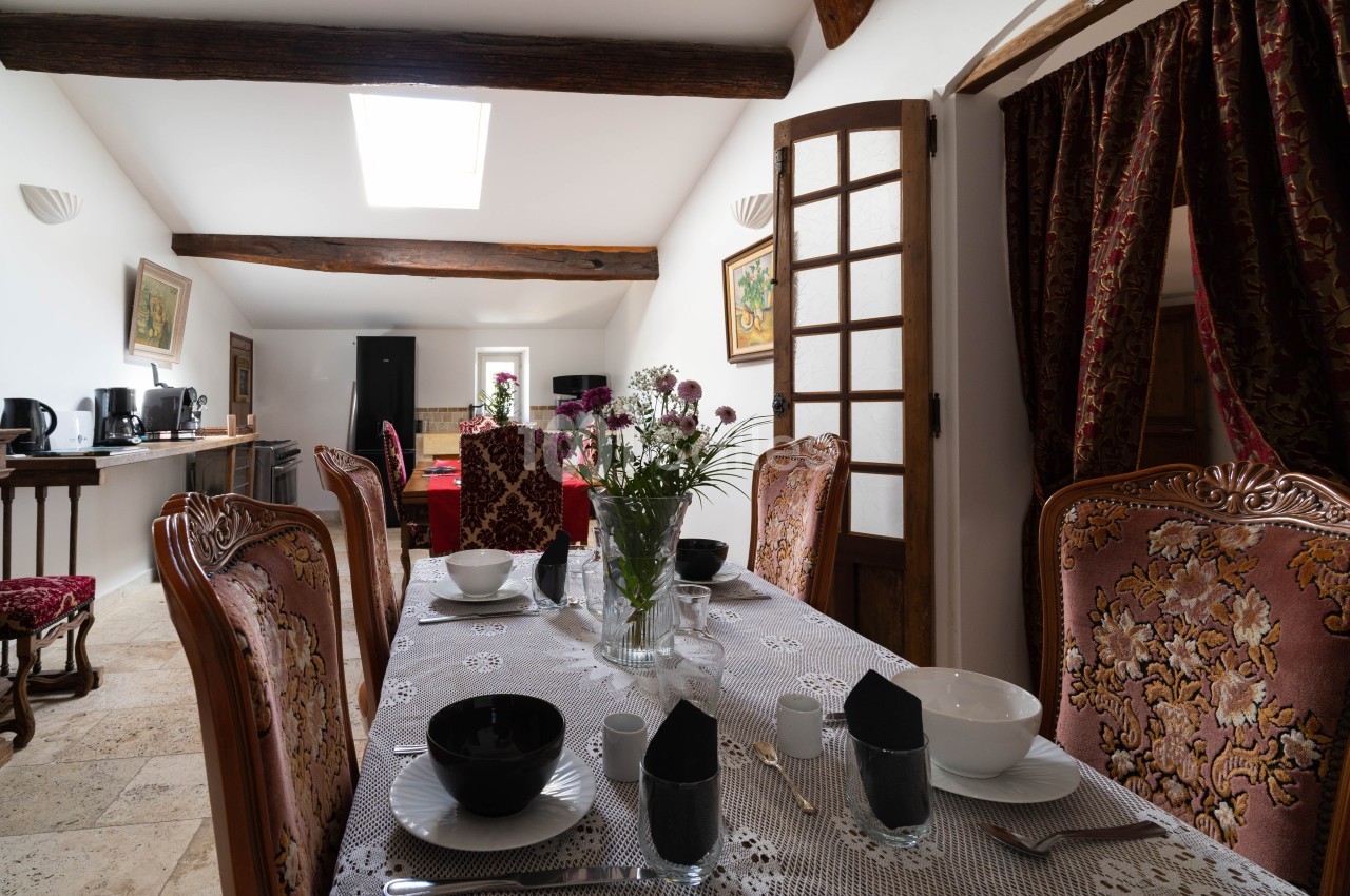 Salle à manger lumineuse avec table dressée, chaises ornées, poutres apparentes et vue sur une cuisine équipée.