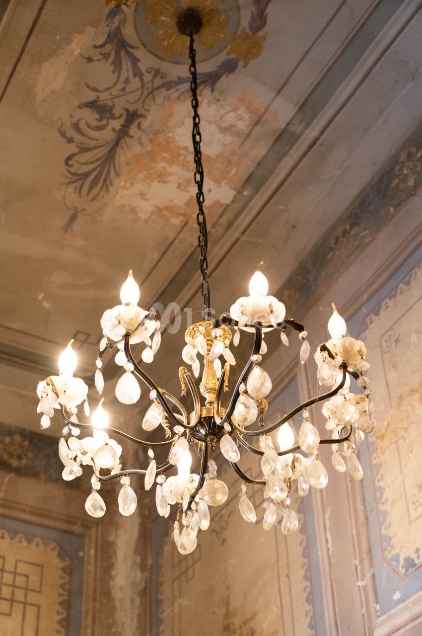 Lustre ancien en métal noir et doré avec pampilles en cristal, suspendu dans une pièce au plafond décoré et vieilli.