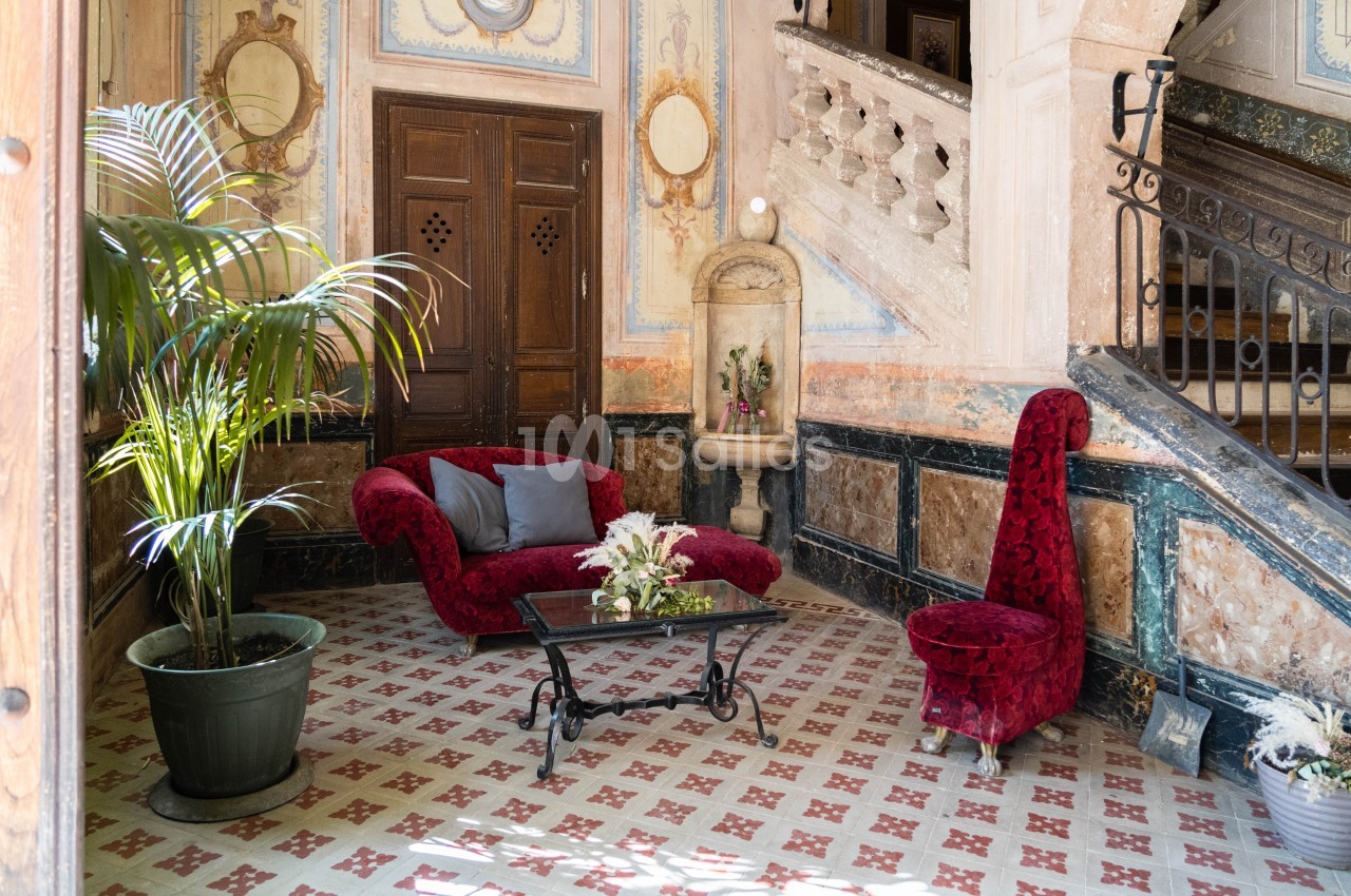 Salon avec fauteuils rouges, table basse en verre, plante en pot et fontaine décorative dans un intérieur ancien.