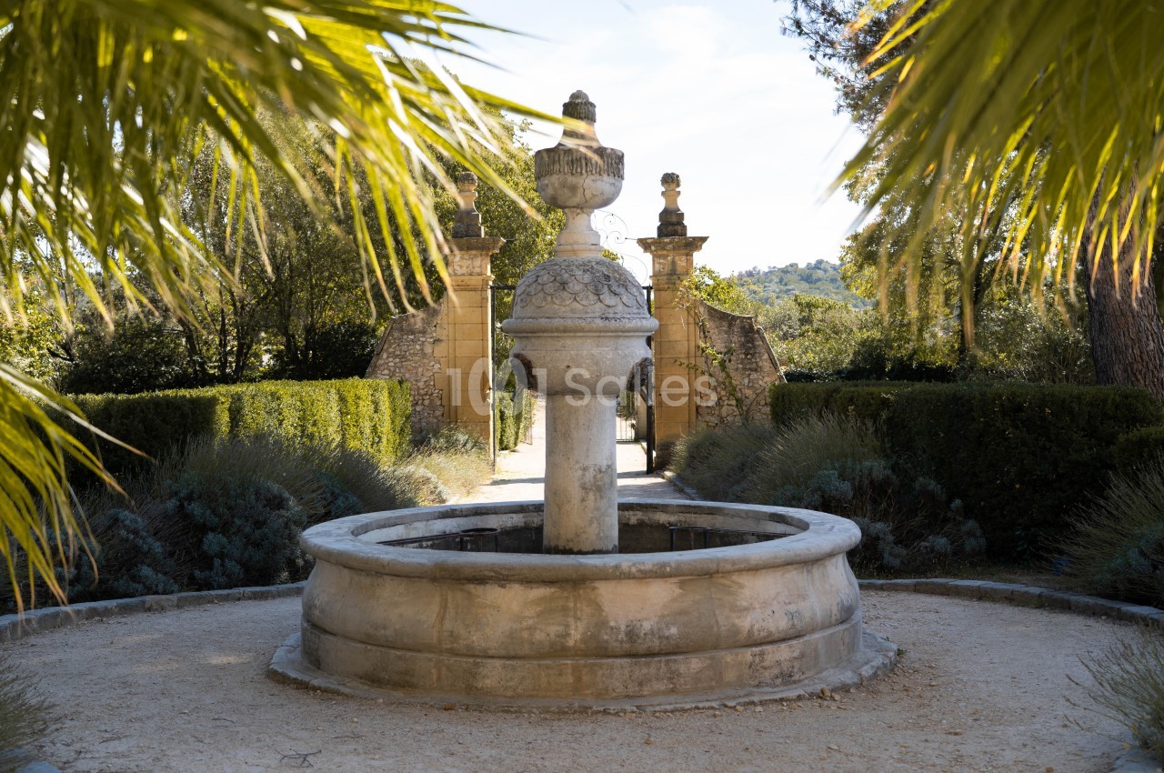 Fontaine en pierre entourée de végétation, avec une allée menant à un portail en arrière-plan.