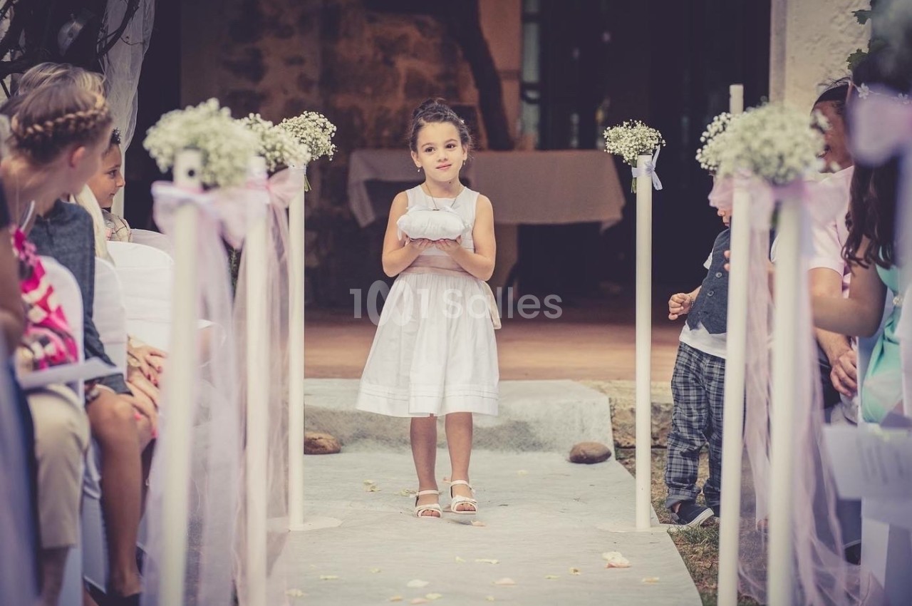 Une jeune fille en robe blanche porte un coussin sur une allée décorée lors d'une cérémonie en plein air.