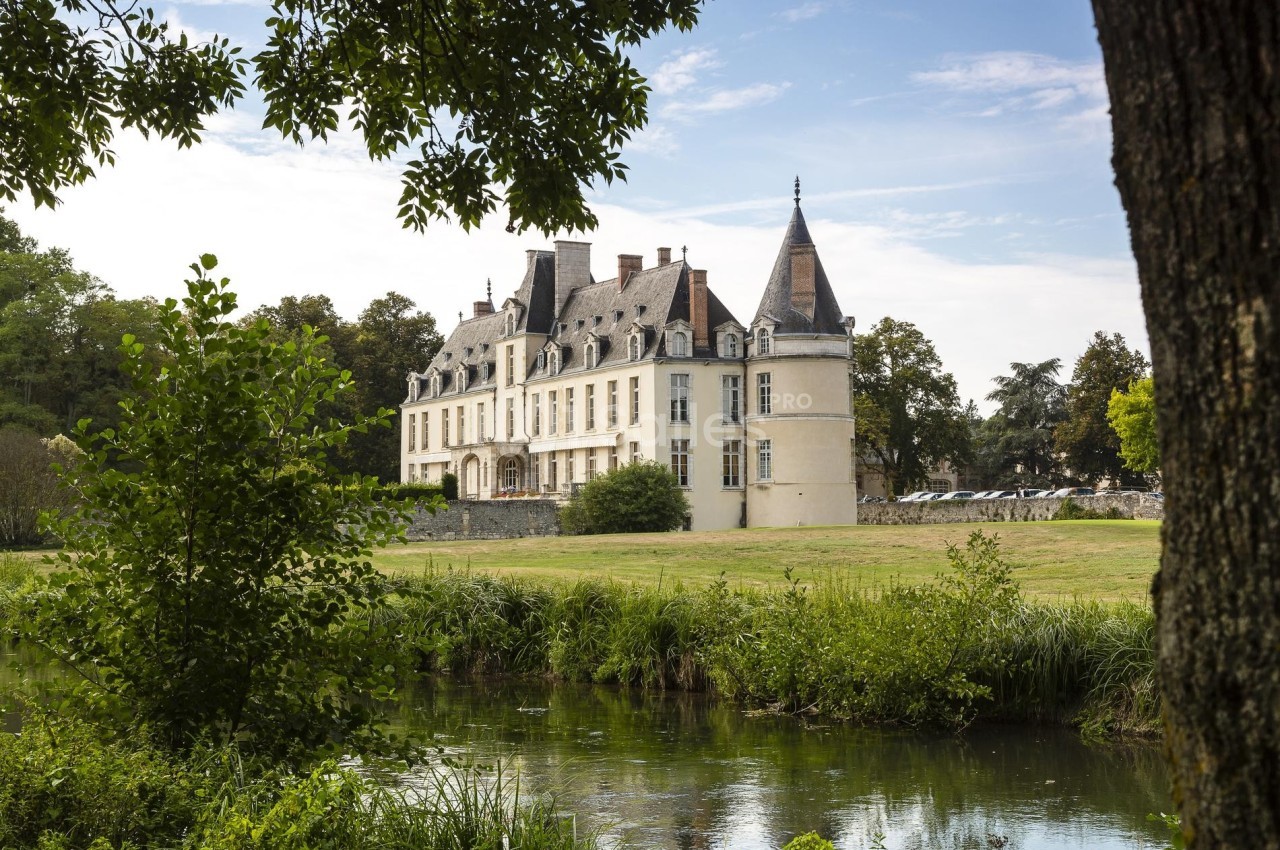 Château entouré de verdure, avec un plan d'eau au premier plan et un ciel partiellement dégagé en arrière-plan.