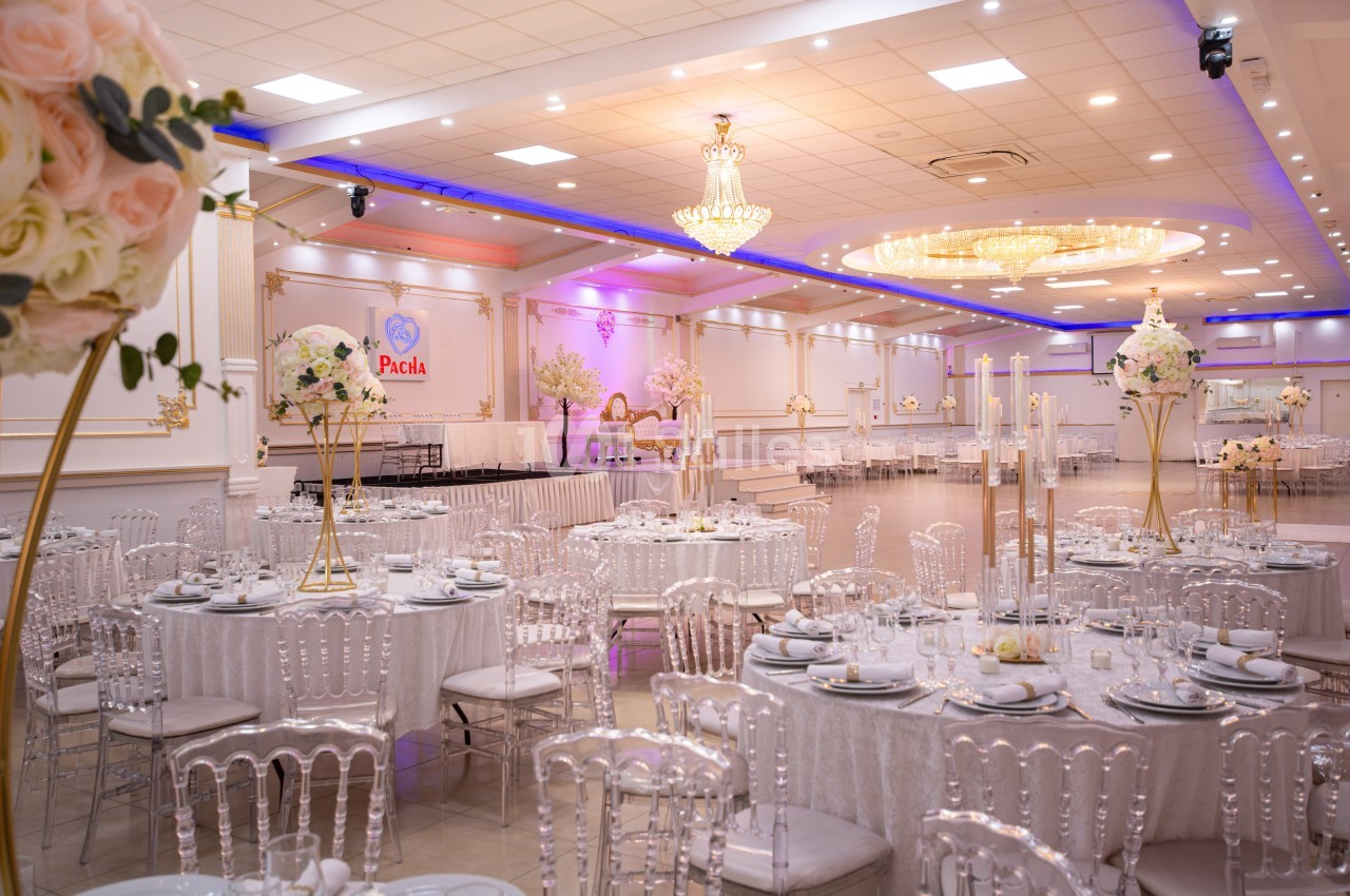 Salle de réception élégante avec tables rondes dressées, chaises transparentes et décorations florales raffinées.
