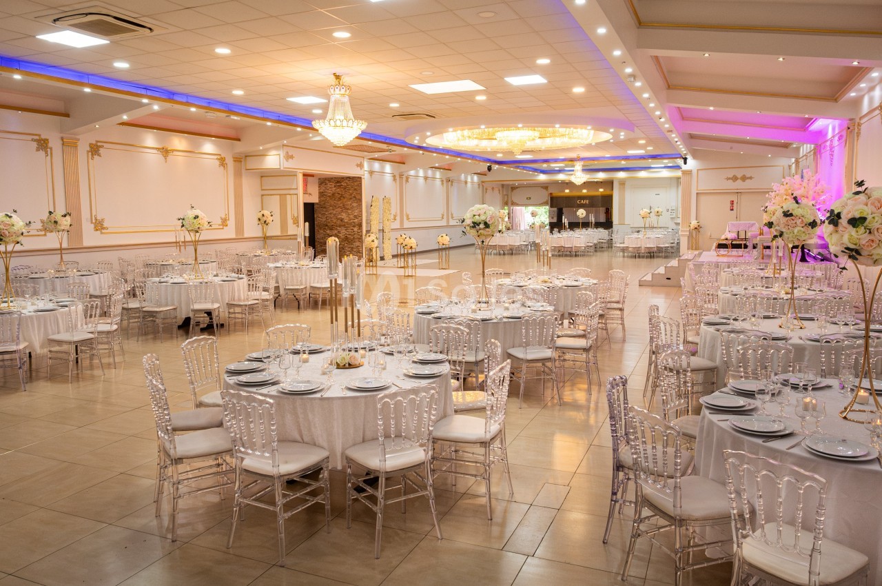 Salle de réception élégante avec tables rondes dressées, chaises transparentes et décoration florale raffinée.