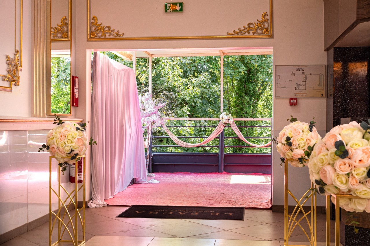 Entrée décorée avec des fleurs blanches et roses, donnant sur une terrasse ornée de drapés et d'arbres en arrière-plan.