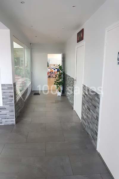 Couloir lumineux avec carrelage gris, murs clairs, plantes décoratives et vue sur une salle en arrière-plan.