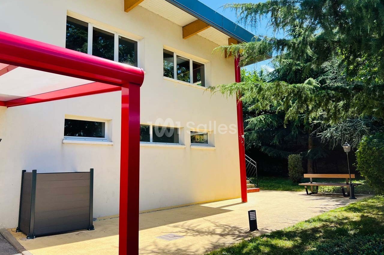 Façade d'un bâtiment moderne avec des fenêtres, un auvent rouge et un espace extérieur arboré avec banc.