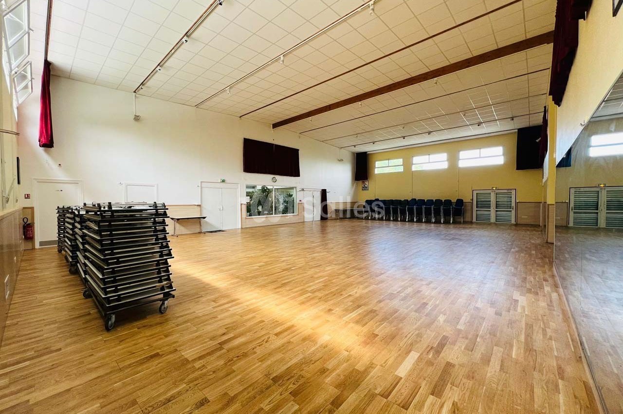 Salle spacieuse avec parquet, chaises empilées sur le côté et tables rangées sur des chariots, éclairée par des fenêtres.