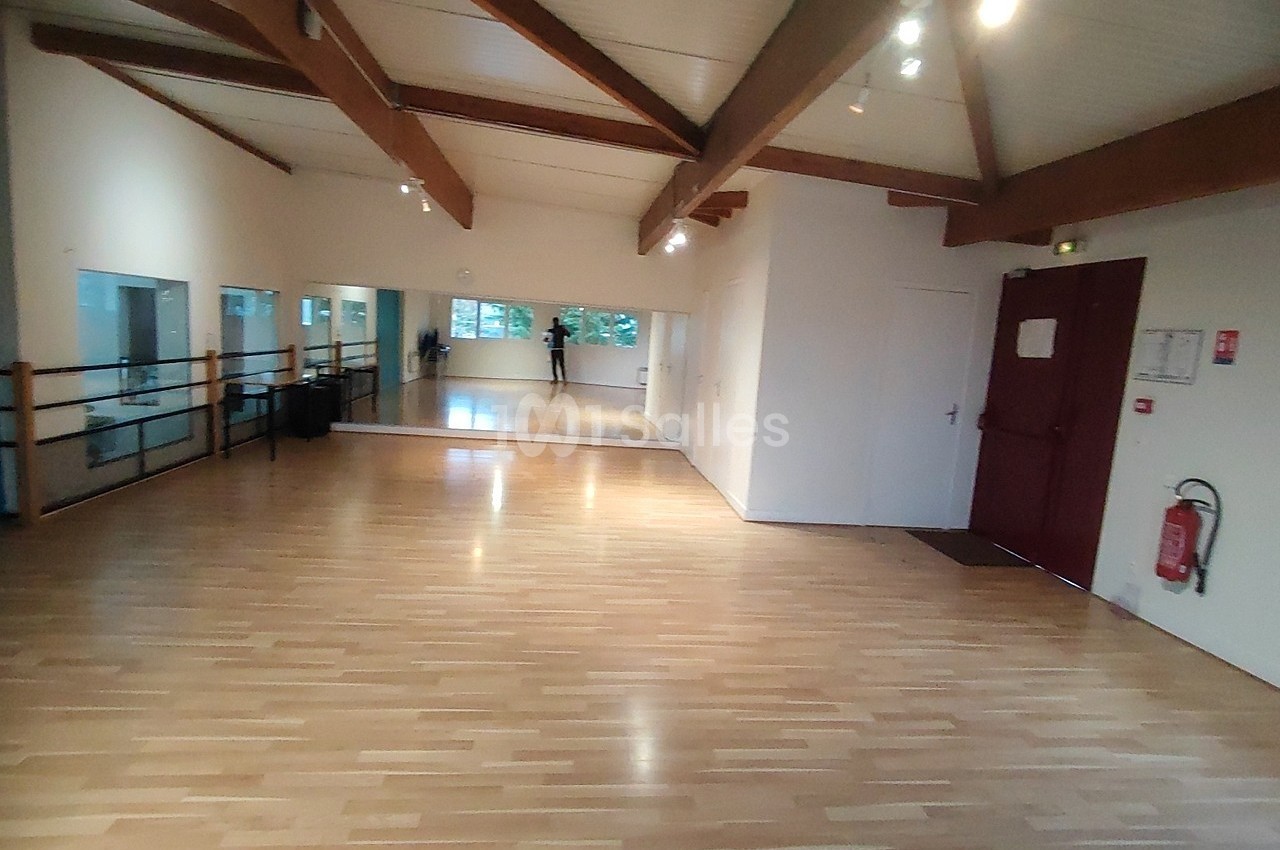 Salle lumineuse avec parquet en bois, miroirs sur un mur, barres de danse et poutres apparentes au plafond.