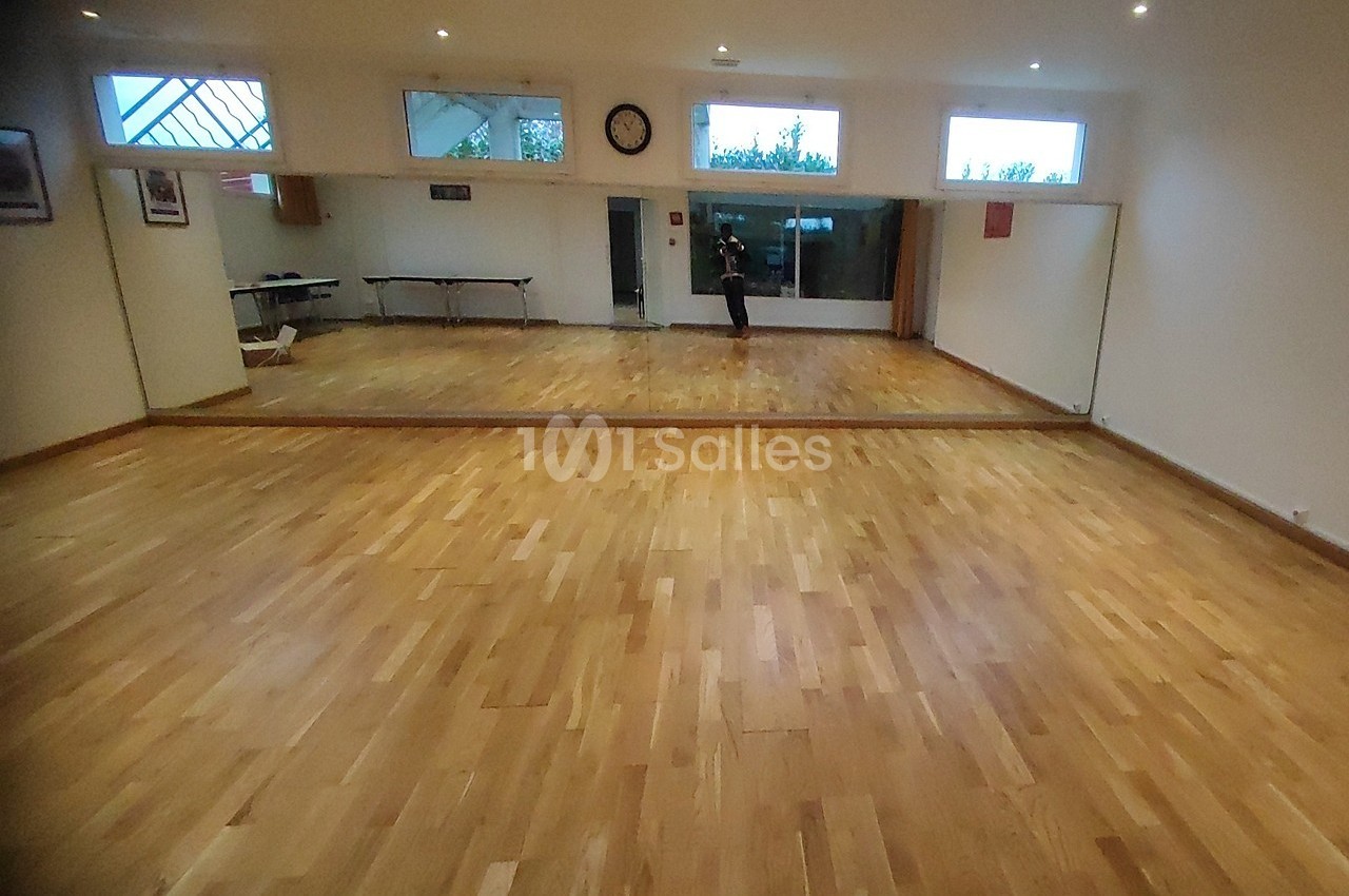 Salle avec parquet en bois clair, grand miroir mural, fenêtres en hauteur et mobilier minimaliste en arrière-plan.