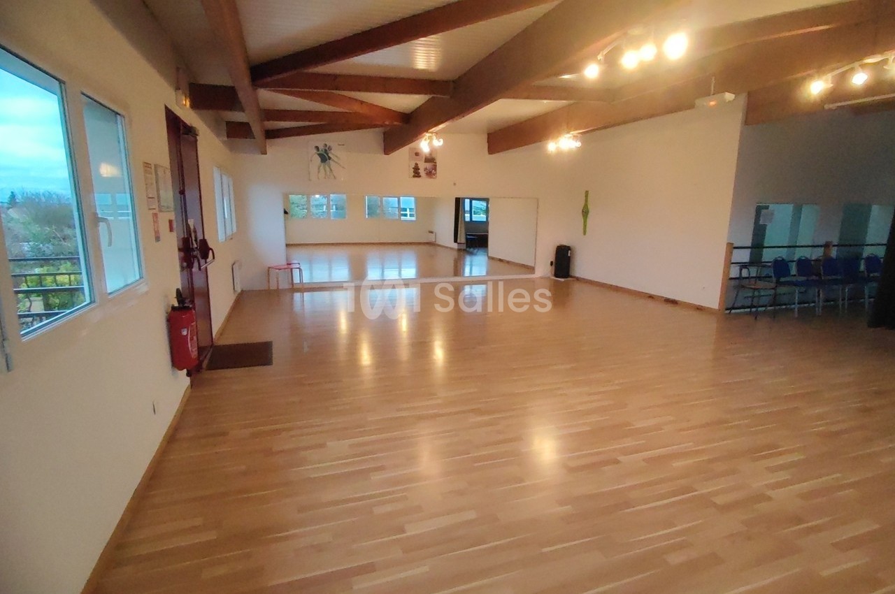 Salle lumineuse avec parquet en bois, murs blancs, grandes fenêtres et plafond avec poutres apparentes.