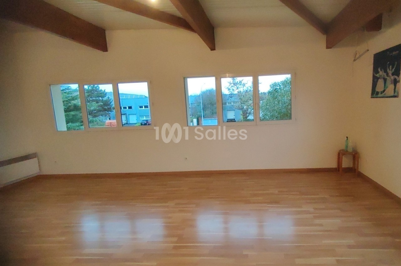 Salle vide avec parquet en bois, murs clairs, grandes fenêtres laissant entrer la lumière naturelle et poutres apparentes.
