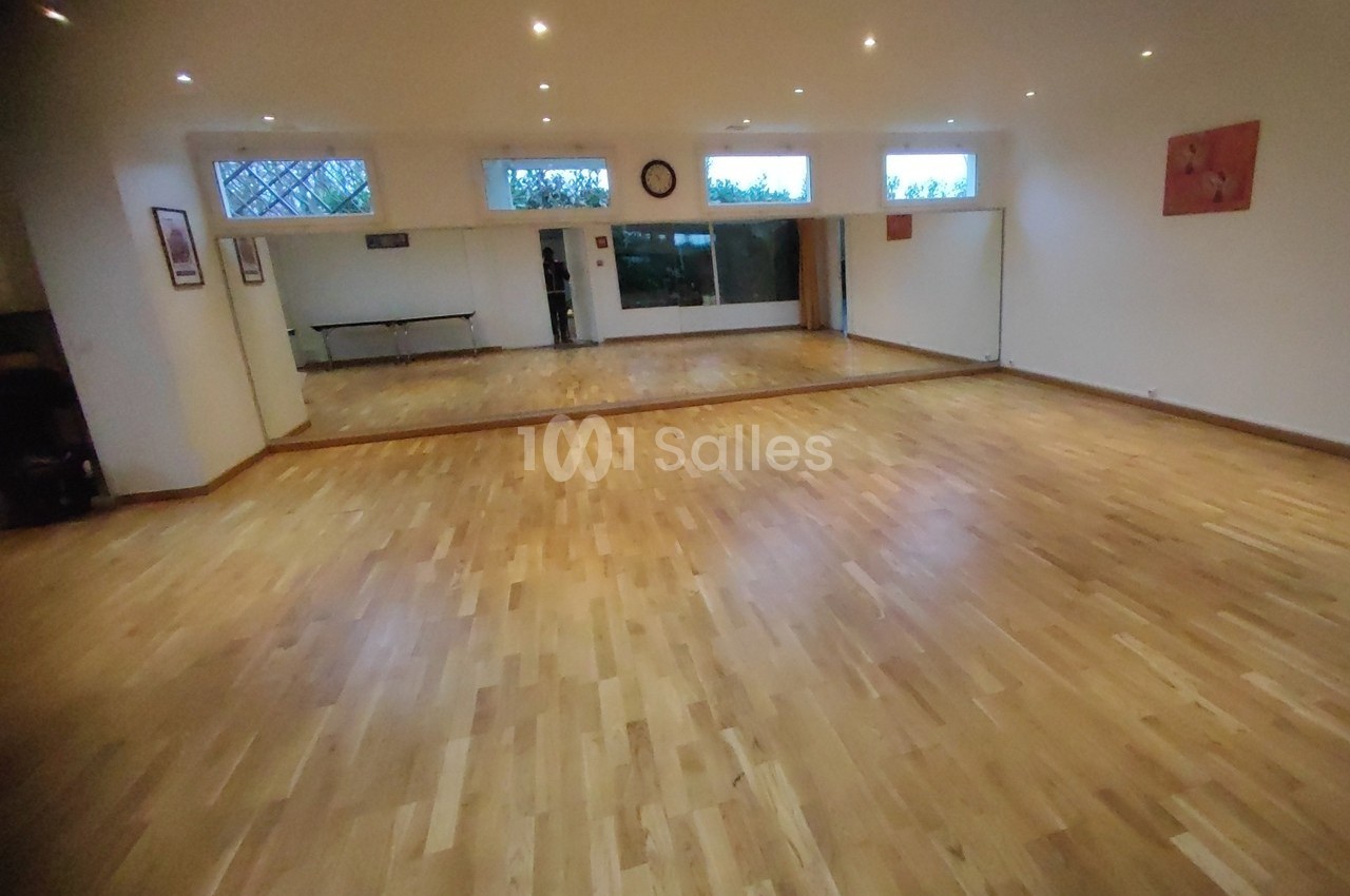 Salle lumineuse avec parquet en bois clair, murs blancs, miroirs sur un mur et fenêtres en hauteur.