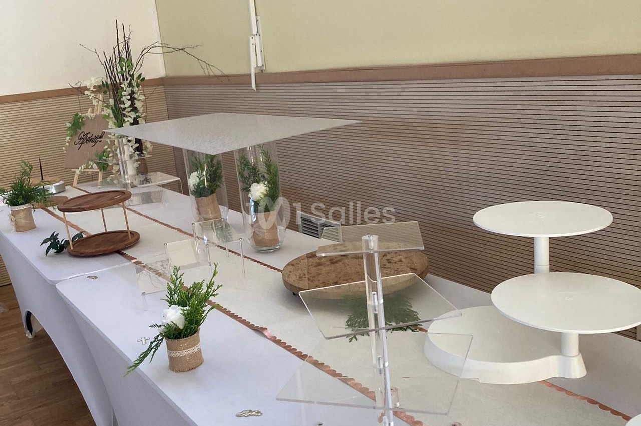 Table décorée pour un buffet, avec supports vides, plantes en pot, branches et nappes blanches dans une salle lumineuse.