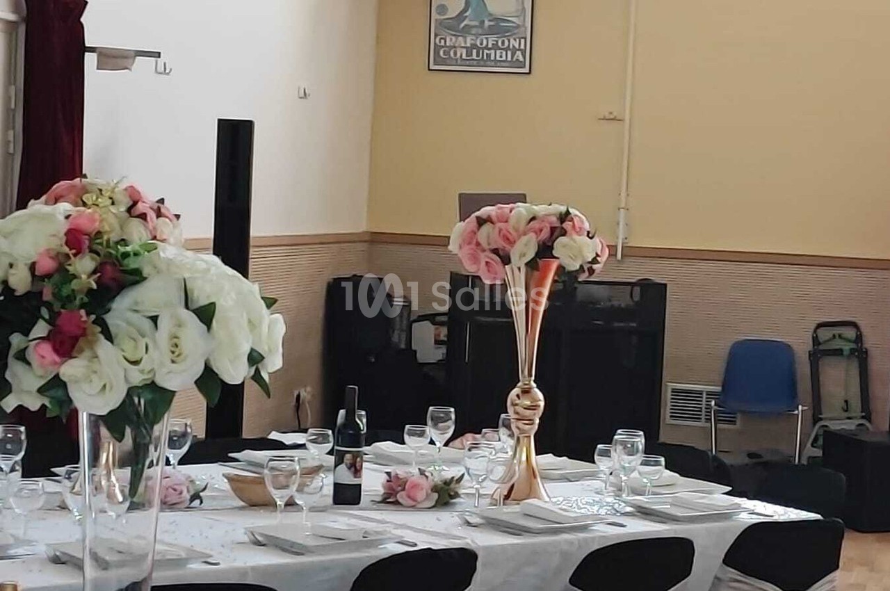 Table dressée pour un événement avec nappes blanches, verres, assiettes et bouquets de fleurs en centre de table.