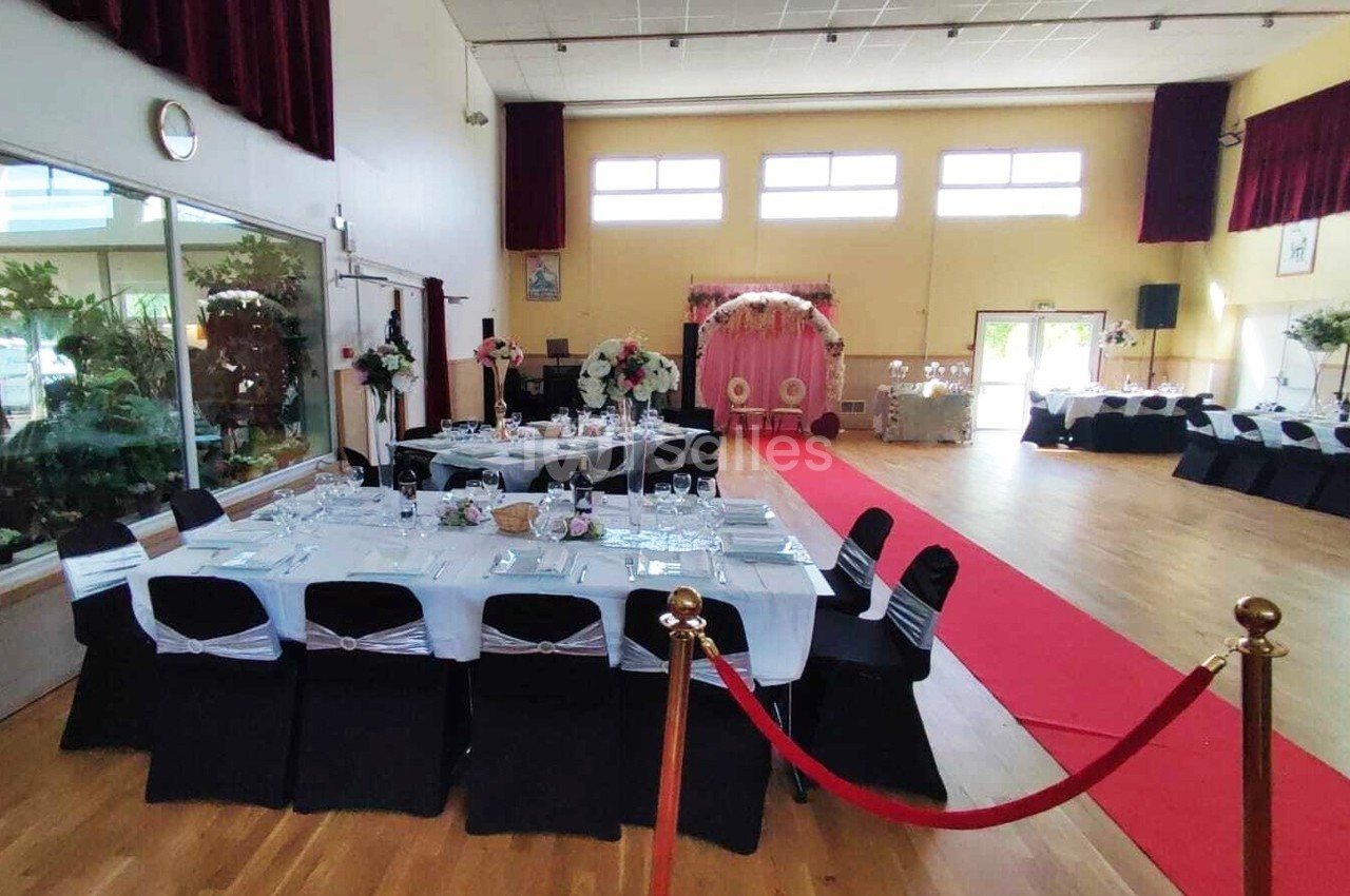 Salle de réception décorée pour un événement, avec tables dressées, allée rouge et arche fleurie au fond.