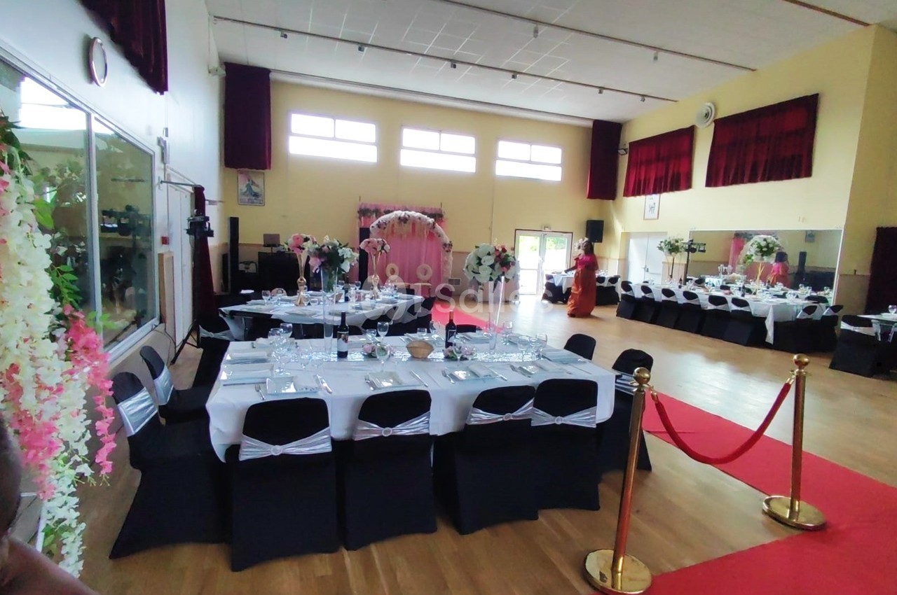 Salle décorée pour un événement avec tables dressées, chaises noires, fleurs et tapis rouge menant à une arche florale.