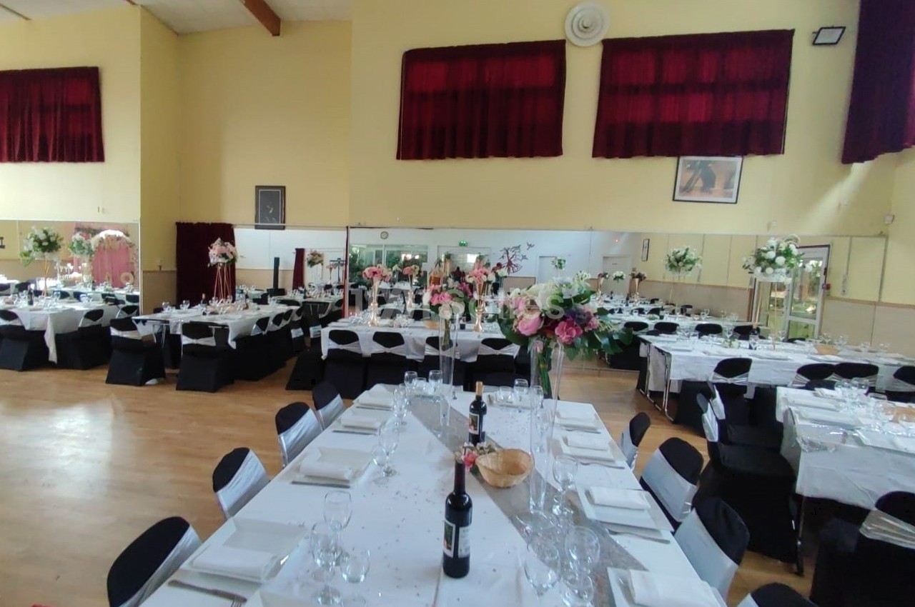 Salle de réception décorée avec des tables dressées, nappes blanches, chaises noires et arrangements floraux.