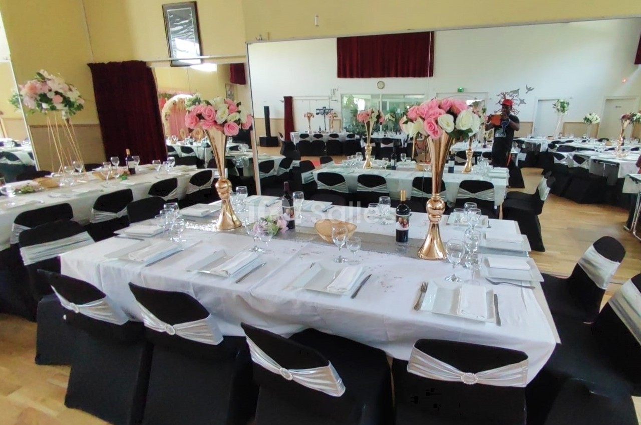 Salle de réception décorée avec des tables dressées, nappes blanches, chaises noires et arrangements floraux.