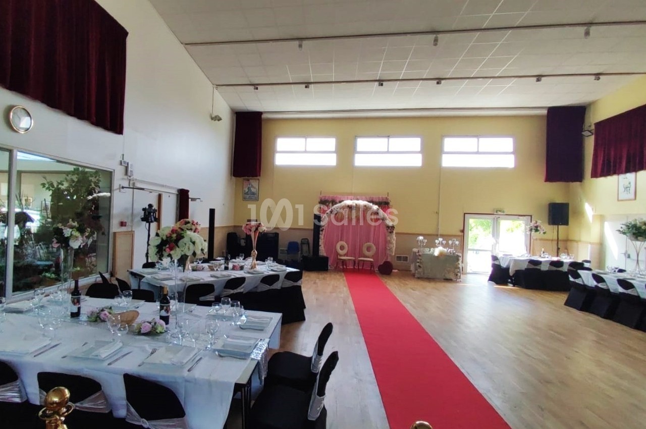Salle de réception décorée pour un événement, avec tables dressées, tapis rouge et arche florale au fond.