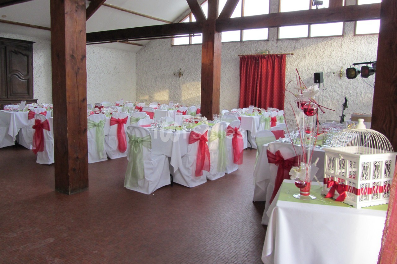 Salle de réception décorée avec des tables nappées de blanc, ornées de nœuds rouges et verts, sous une charpente en bois.
