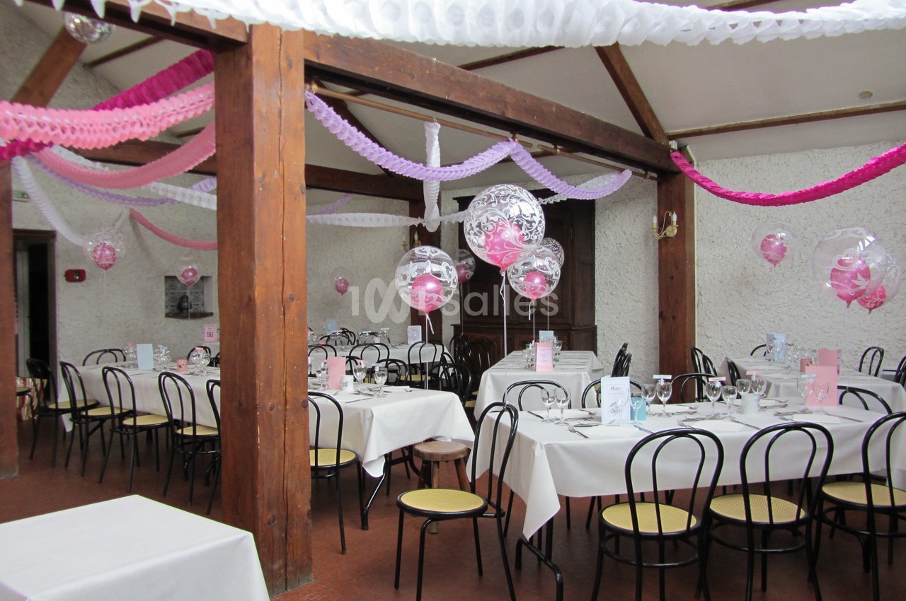Salle décorée pour une réception avec guirlandes colorées, ballons transparents et tables dressées avec nappes blanches.