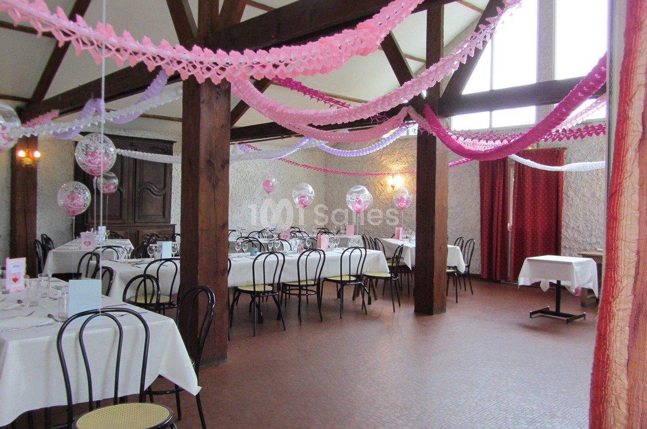 Salle de réception décorée avec des guirlandes roses et violettes, tables dressées avec nappes blanches et chaises noires.