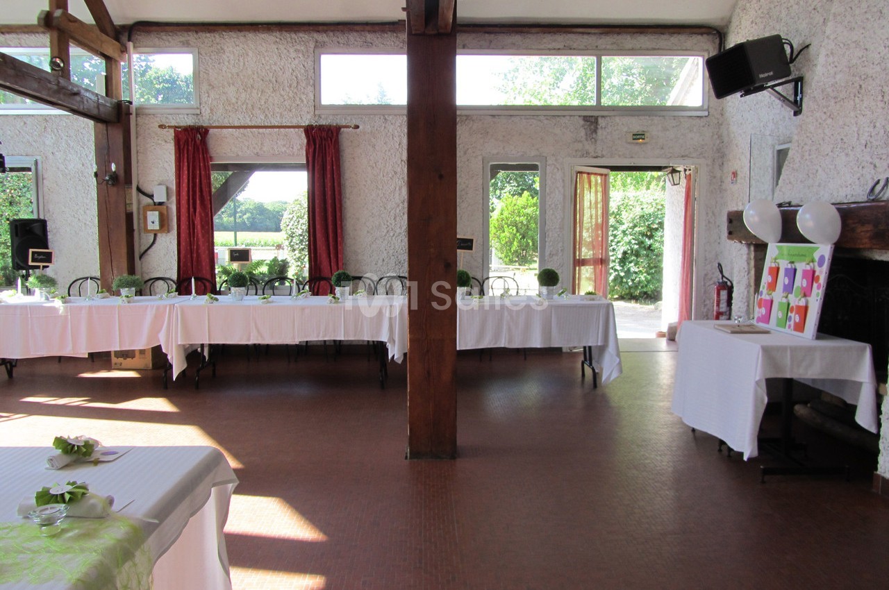 Salle lumineuse avec de grandes fenêtres, tables dressées avec nappes blanches et vue sur un jardin verdoyant.