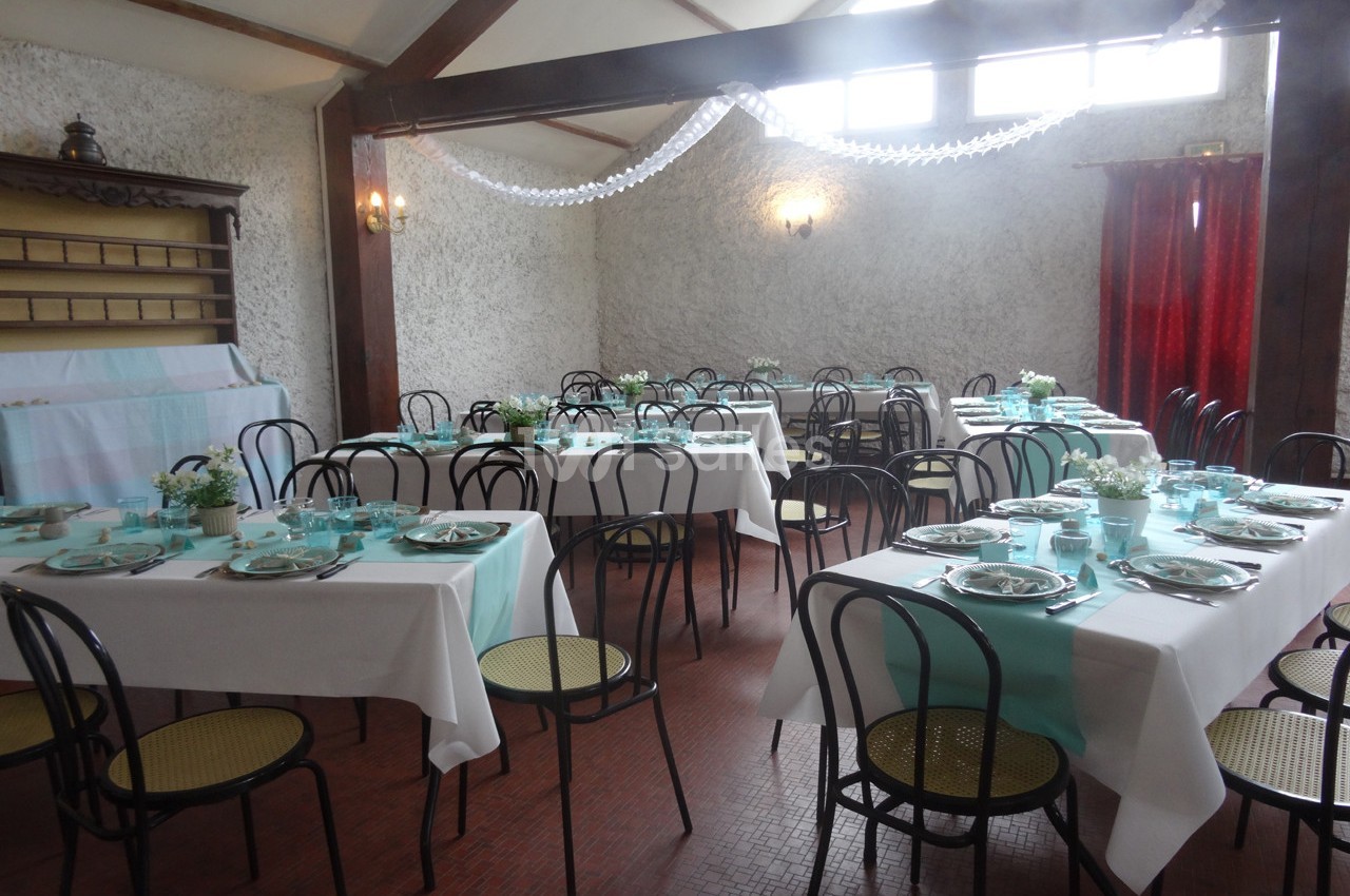 Salle de réception aménagée avec des tables dressées, nappes blanches et décorations bleu pastel, éclairée par des fenêtres.