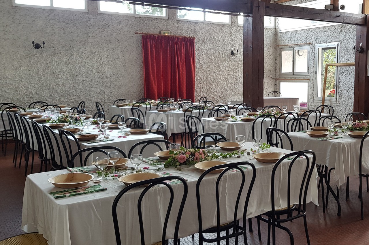 Salle de réception avec tables dressées, nappes blanches, chaises noires et décoration florale au centre des tables.