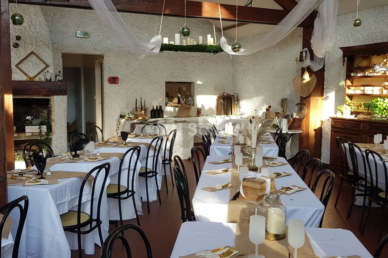Salle de réception décorée avec des tables dressées, nappes blanches, chaises noires et éléments naturels.