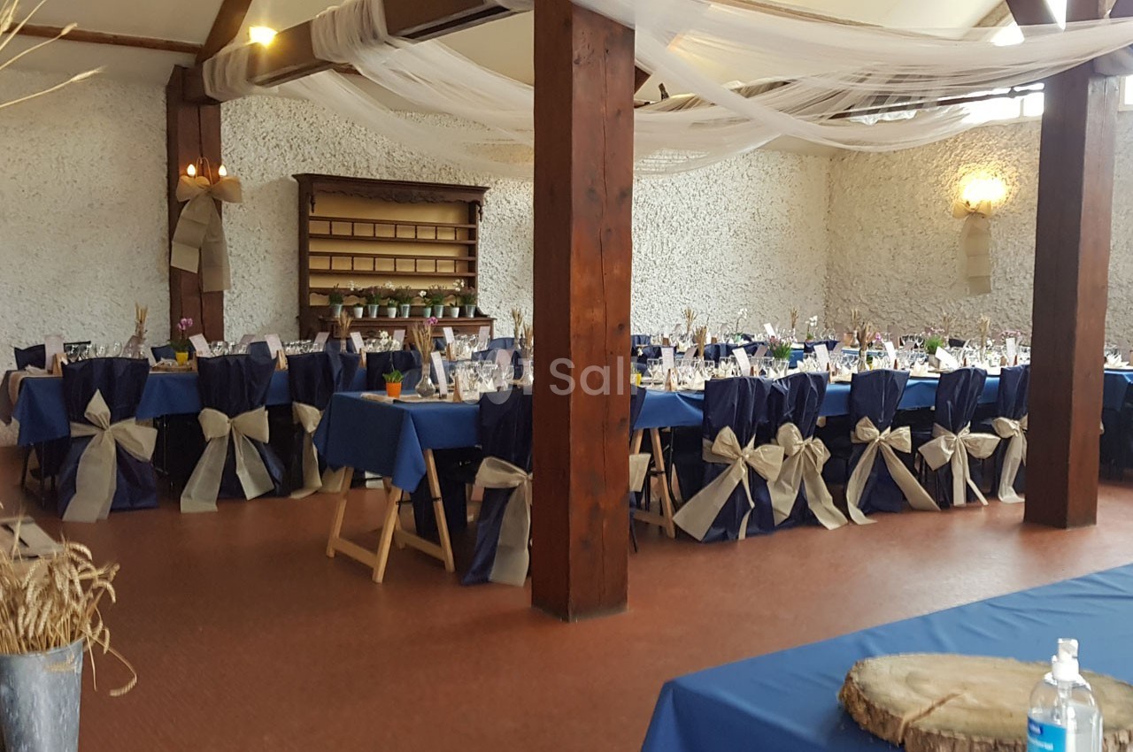 Salle de réception décorée avec des tables dressées, nappes bleues et chaises ornées de nœuds beiges.