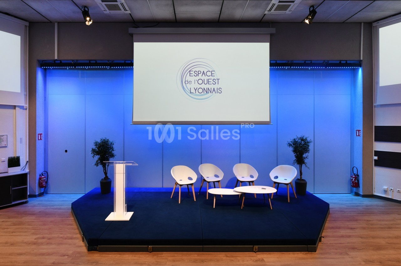 Salle de conférence moderne avec scène, pupitre, quatre chaises blanches et écran projetant un logo.