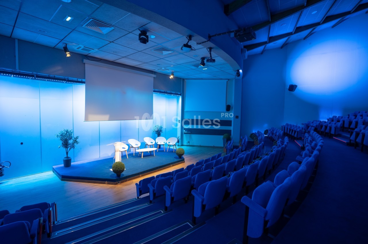 Salle de conférence vide avec des sièges alignés, une scène éclairée, des chaises blanches et un écran de projection.