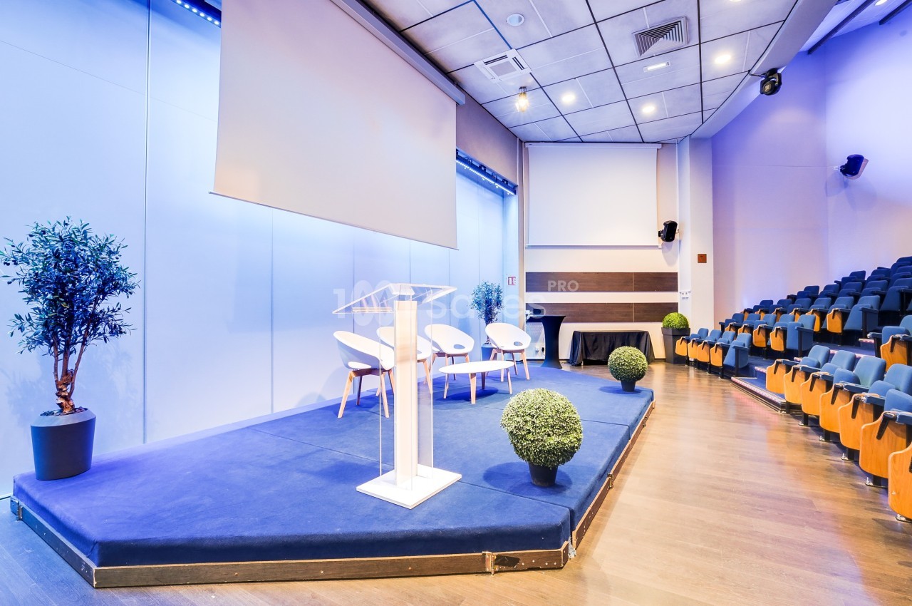 Salle de conférence moderne avec estrade, pupitre transparent, chaises blanches et rangées de sièges vides.