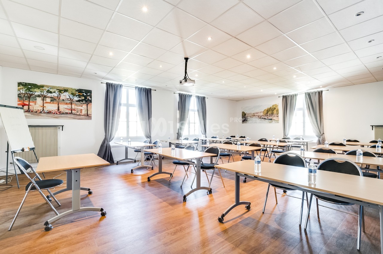 Salle de réunion lumineuse avec tables, chaises, projecteur au plafond et grandes fenêtres avec rideaux.