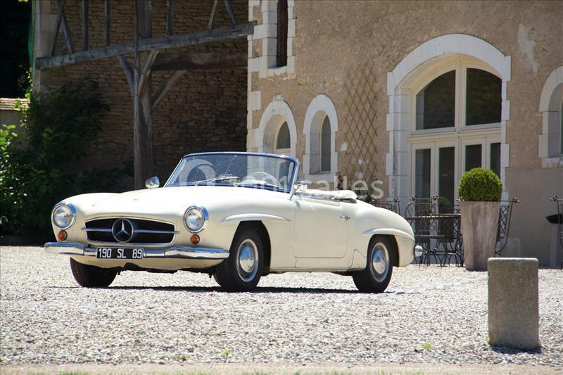 Voiture ancienne décapotable blanche stationnée devant une maison en pierre avec grandes fenêtres et terrasse.