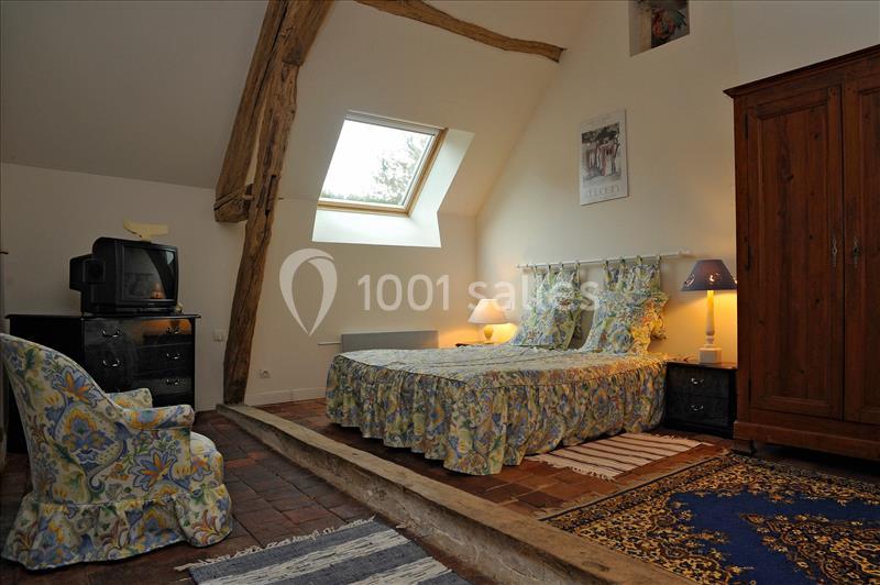 Chambre mansardée avec lit double, poutres apparentes, armoire en bois, télévision et fauteuil sur sol en tomettes.
