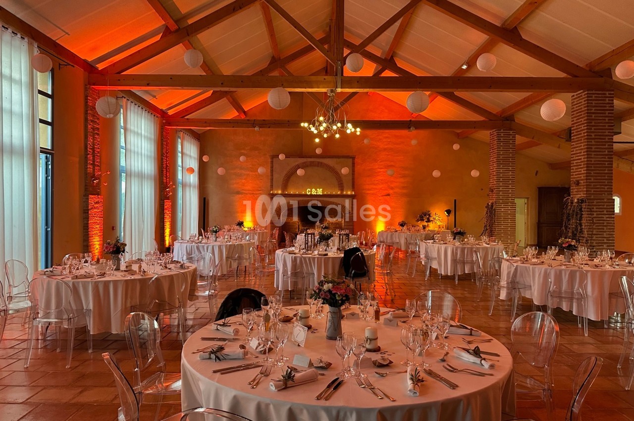 Salle de réception décorée avec des tables rondes dressées, éclairage chaleureux et poutres apparentes.