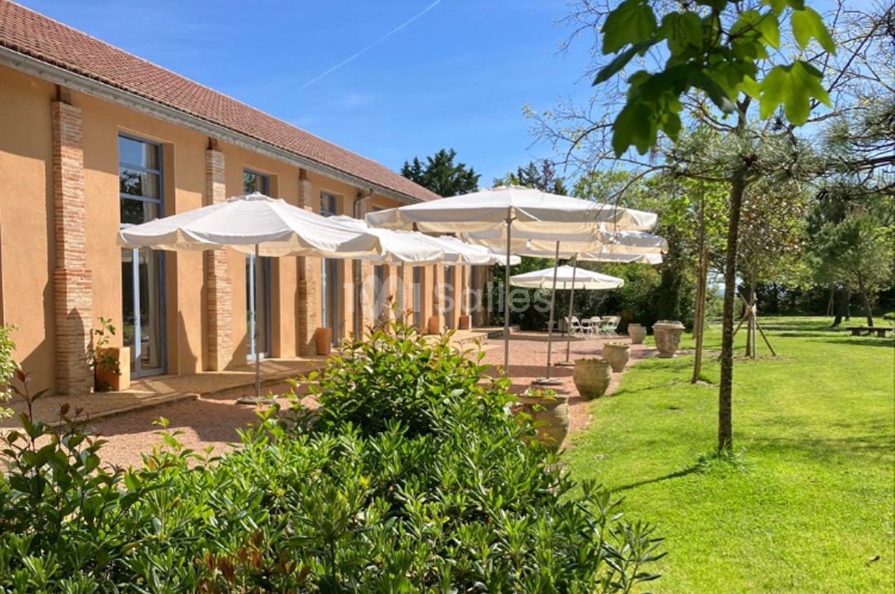 Terrasse ensoleillée avec parasols blancs devant un bâtiment ocre, entourée de verdure et d'arbres.