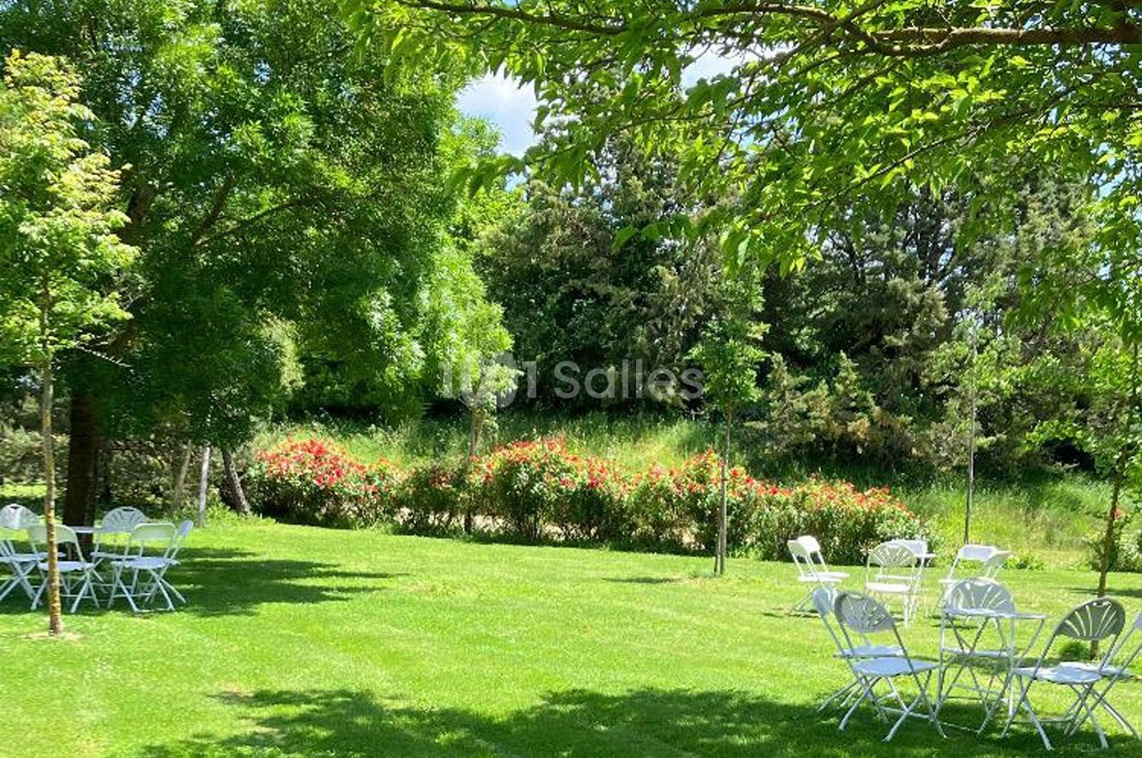 Chaises et tables blanches disposées sur une pelouse verdoyante entourée d'arbres et de buissons fleuris.