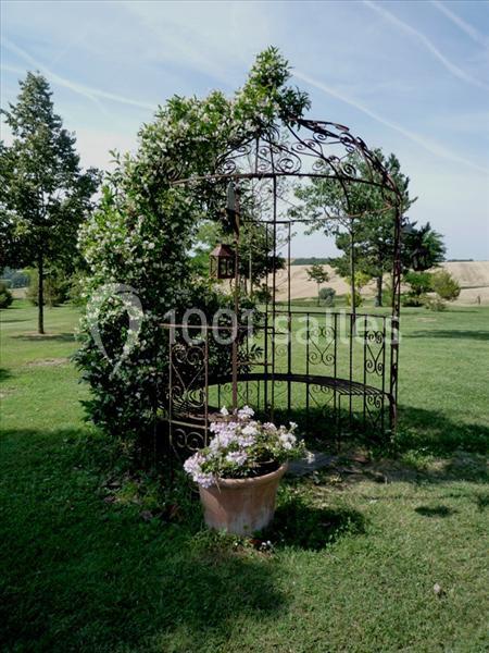 Arche en fer forgé recouverte de végétation, entourée d'herbe, avec un pot de fleurs au premier plan.
