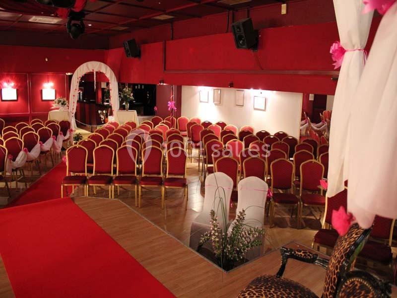 Salle décorée en rouge avec des rangées de chaises alignées, un espace scénique et des arches ornées de fleurs blanches.