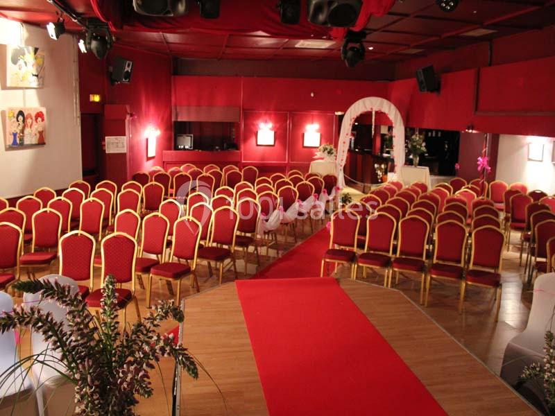 Salle de réception décorée en rouge avec des rangées de chaises alignées et une arche fleurie au fond.
