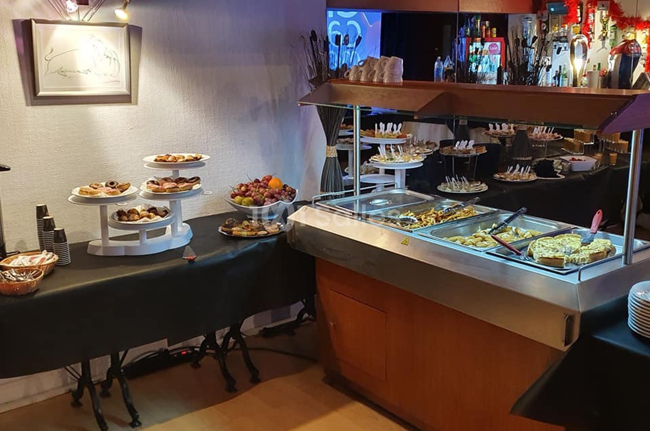 Buffet avec plats chauds, desserts variés, fruits et vaisselle disposés sur des tables dans un espace éclairé.