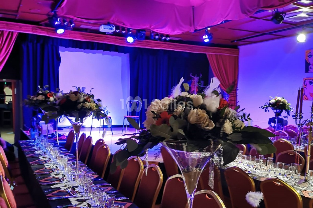 Salle de réception décorée avec des tables alignées, des compositions florales et une scène éclairée en arrière-plan.